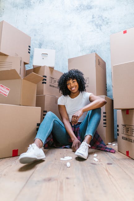 A woman with curly black hair, wearing a white t-shirt, blue jeans, and white sneakers, is sitting on a wooden floor surrounded by numerous cardboard moving boxes of varying sizes, some with labels an