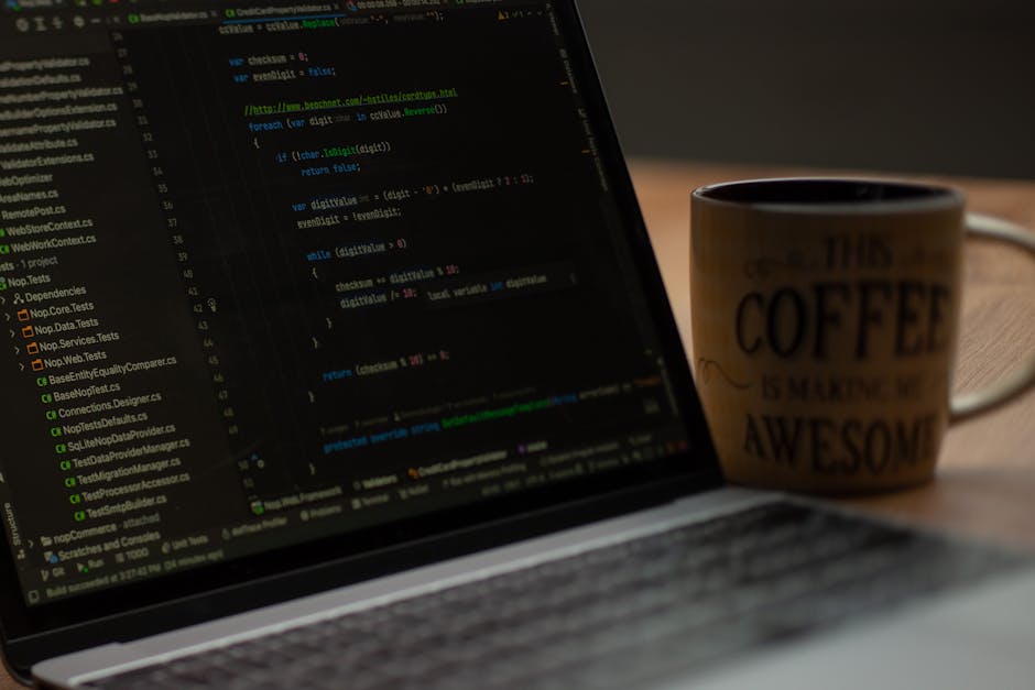 A close-up image of a person's hand operating a laptop computer on a wooden surface, with visible code displayed on the screen. The individual is wearing a dark wristwatch, and the laptop has a silver